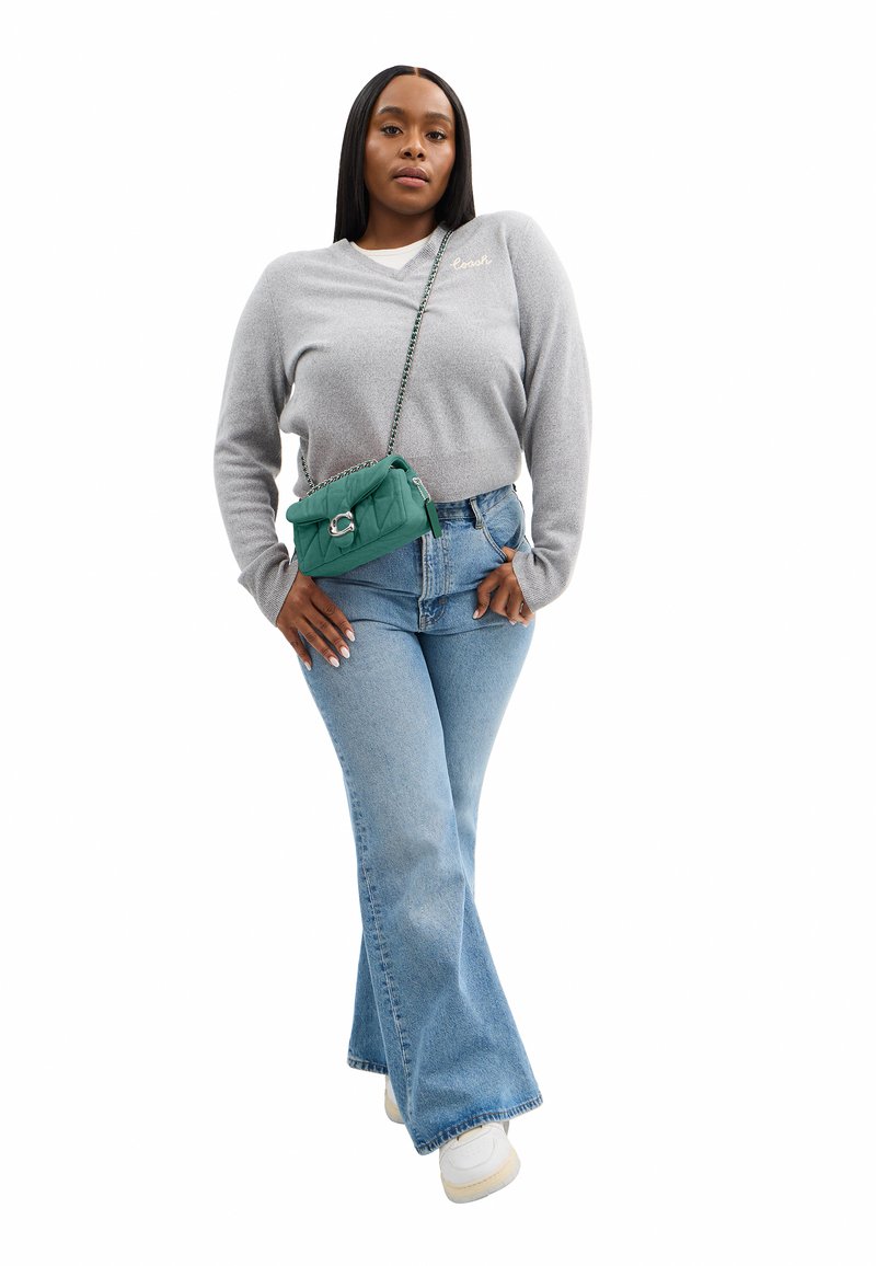 Kvinde iført en lysegrå sweater og blå jeans står med den ene hånd i lommen og bærer en grøn quiltet crossbodytaske.