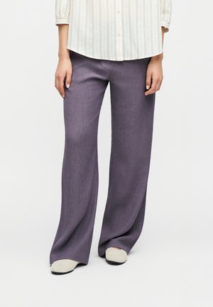 Femme portant un pantalon large violet, des chaussures blanc cassé et une chemise rayée boutonnée rentrée, debout devant un fond uni.