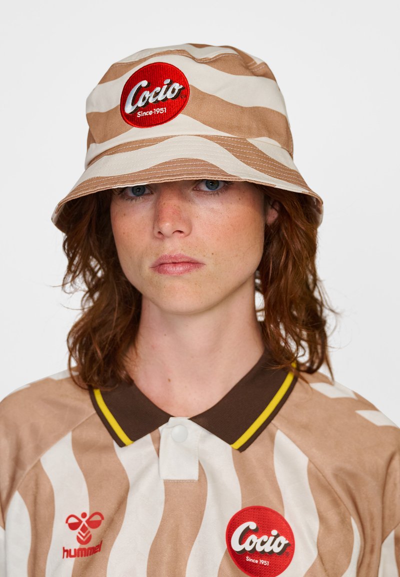 Bucket-hatt med bruna och beige vågiga ränder, med en röd cirkulär logotyp-patch. Matchande mönstrad skjorta med krage och gula detaljer.