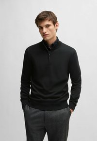 Black zip-neck sweater in lightweight knit fabric with a fitted design. Features long sleeves and ribbed cuffs and hem, paired with gray trousers.