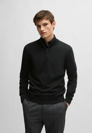 Svart zip-neck jumper i lätt stickad kvalitet med en figurnära design. Har långa ärmar och ribbade muddar och fåll, i kombination med gråa byxor.