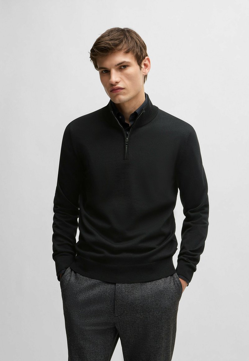Black zip-neck sweater in lightweight knit fabric with a fitted design. Features long sleeves and ribbed cuffs and hem, paired with gray trousers.