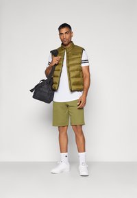 Olivengrüner Puffer-Weste über einem weißen T-Shirt mit schwarzen Streifen, kombiniert mit grünen Shorts, weißen Sneakern und einer schwarzen Sporttasche.