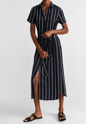 Femme portant une robe midi à manches courtes à rayures verticales noires et blanches, avec une ceinture nouée à la taille et des sandales noires ouvertes.