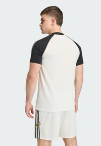 Λευκό πουκάμισο με κοντά μανίκια και μαύρα ράγκλαν μανίκια. Διαθέτει ομαλή υφή υφάσματος. Συνδυάζεται με λευκά σορτς που φέρουν μαύρες ρίγες και λογότυπο.