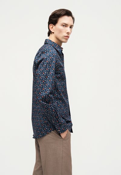 Νέος άνδρας με μπλε πουκάμισο με μικρά πολύχρωμα γεωμετρικά σχήματα και μπεζ παντελόνι, όρθιος με τα χέρια στις τσέπες, κοιτάζοντας προς τα πλάγια.