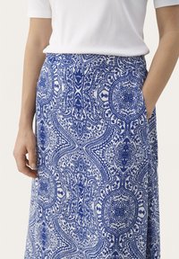 Frau trägt einen Rock mit blau-weißen Muster, hat ihre rechte Hand in der Tasche und ein weißes, geripptes Kurzarmoberteil.