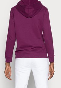 Fialová mikina s kapucí, klokáním klokáním, žebrovanými manžetami a lemem, s kapucí se stahovací šňůrkou, nošená přes bílé kalhoty.