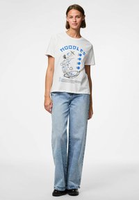 Camiseta blanca con un diseño gráfico azul de fideos en un tazón, combinada con jeans de pierna ancha de color azul claro y zapatos negros.