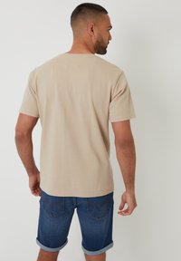T-shirt beige a maniche corte in tessuto texture, con dettaglio sulla spalla quadrato. Abbinato a pantaloni jeans a pinces con risvolto in un lavaggio blu scuro.