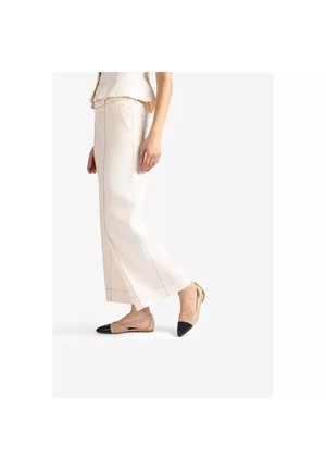 Femme portant un pantalon large beige avec des ballerines beige à bouts noirs, debout de profil sur fond blanc.