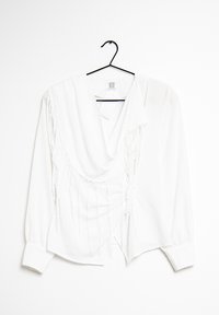 Vit långärmad blus med draperad halsringning, som har vertikala fransdetaljer på ena sidan och knäppta manschetter. Hänger från en svart galge.