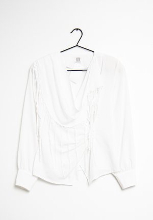 Blouse blanche à manches longues avec un col drapé, présentant des détails de franges verticales sur un côté et des poignets à boutons. Accrochée à un cintre noir.