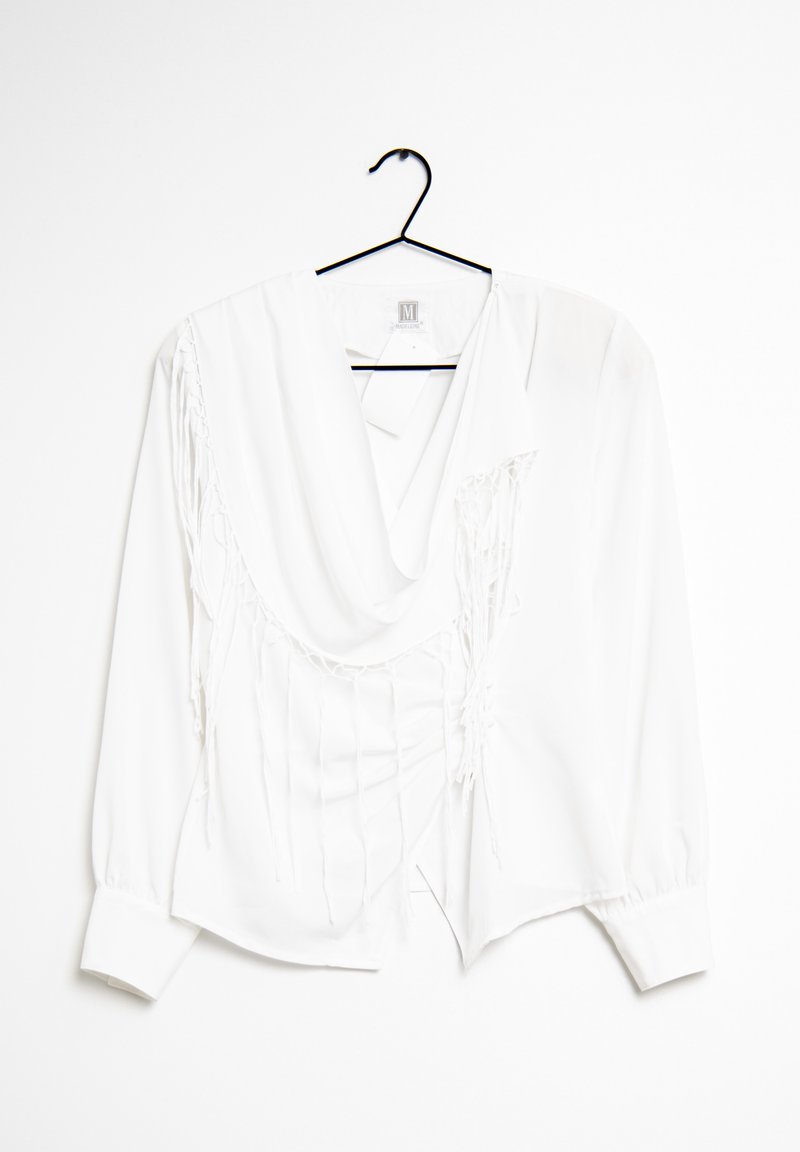 Vit långärmad blus med draperad halsringning, som har vertikala fransdetaljer på ena sidan och knäppta manschetter. Hänger från en svart galge.