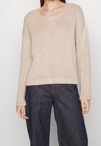 Beżowy gruby sweter dzianinowy z dekoltem w kształcie V noszony z ciemnymi jeansami typu wide-leg przez stojącą osobę na jednolitym tle.