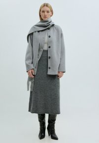 Lichtgrijze wollen jas met grote knopen, gecombineerd met een donkergrijze, knie-lange rok en kniehoge zwarte leren laarzen. Sjaal hangt elegant.