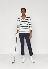 Fehér és sötétkék csíkos V nyakú pulóver, bordás textúrával, sötétkék nadrággal és fehér golfcipővel párosítva. Az egyik kezében golfütőt tart.