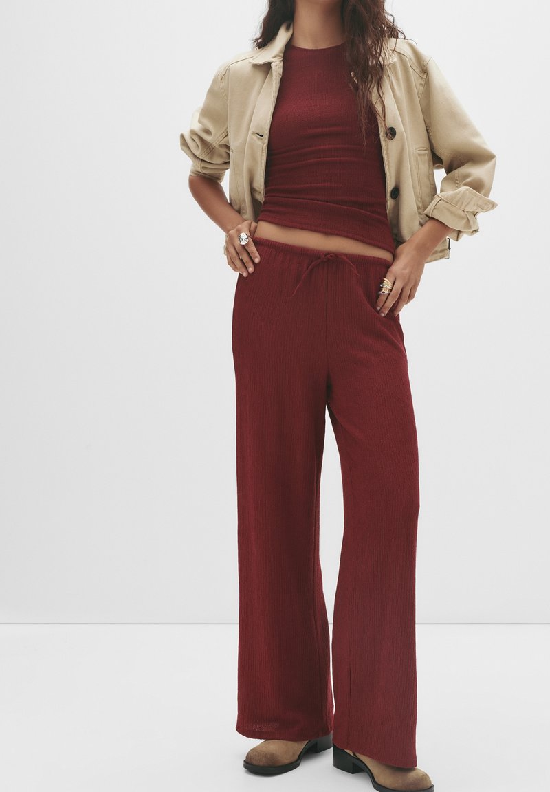 Top a costine bordeaux e pantaloni a gamba larga abbinati a un giubbotto beige corto. Scarpe con tacco marroni e anelli sovrapposti completano l'outfit.