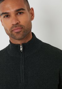 Donkergroene ritssluitingtrui met een hoge kraag, voorzien van een gemêleerd breisel en een zichtbare zilveren ritssluiting bij de hals.