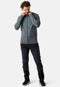 Grijze zip-up jack met een gestructureerd patroon, gecombineerd met zwarte technische broeken met zakken en naden, gedragen met donkere sportschoenen.