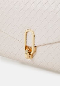 Beige vävd läderväska med en mjuk textur, utrustad med ett spänne och en ring i guldfärgad metall. Närbild av stängningen.