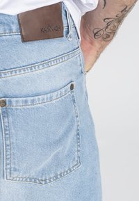 Svijetloplave traperice s smeštim kožnim brendom na stražnjoj strani. Imaju šivenu pregradu s metalnim zakovicama. Glatka tekstura.