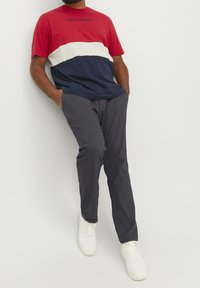 Röd, vit och marinblå randig t-shirt med "JACK & JONES"-logotyp, matchad med mörkgrå byxor och vita sneakers, lutad med händerna i fickorna.