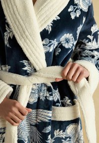 Modrá kvetinová župana s plyšovým bielym golierom a opaskom, s jemnou textúrou a vreckami. Navrhnuté pre pohodlie a teplo.