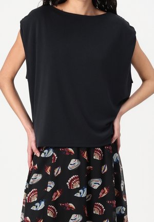 Donna che indossa un top nero senza maniche e una gonna con motivi colorati di conchiglie, le mani appoggiate sui fianchi.