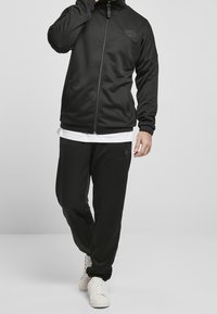 Veste zippée noire et pantalon de survêtement assorti en tissu doux ; présente un logo subtil sur la poitrine et sur le pantalon, avec des poignets et une taille côtelés.