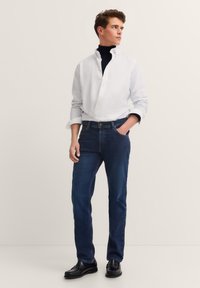 Young man wearing a white button-up shirt over a black turtleneck, dark blue jeans, and black loafers, standing with one hand in pocket.