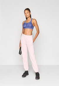 Μωβ αθλητικό σουτιέν με ανοιχτό μπλε λεπτομέρειες και διαμορφωμένα κύπελλα, συνδυασμένο με ανοιχτό ροζ παντελόνια φόρμας με ελαστική μέση και μαύρα αθλητικά παπούτσια.