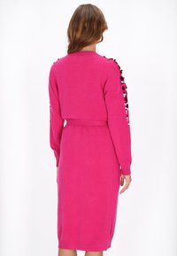 Donna che indossa un abito a maglia rosa brillante con maniche lunghe e cintura, decorato con paillettes sulle maniche, mostrato di schiena.