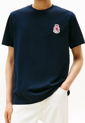 Personne portant un t-shirt bleu marine à manches courtes avec un petit logo blason rouge et blanc sur la poitrine gauche et un pantalon blanc.