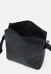 Borsa in pelle nera con una finitura testurizzata, chiusura a zip e una singola tracolla regolabile. L'interno presenta una fodera nera uniforme. Logo "Calvin Klein" visibile.