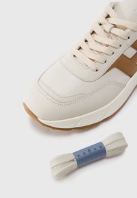 Zapatilla de cuero beige con un acento marrón, suela de goma texturizada y cordones crema. También incluye un par de cordones a juego con una etiqueta azul.