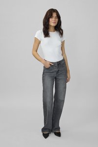 Jauna moteris, dėvinti aptemptą baltą trumpomis rankovėmis palaidinę ir pilkus aukšto liemens, tiesaus kirpimo džinsus, stovinti prieš pilką vientisą foną.