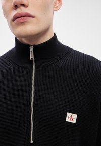 Čierny pletený sveter s vysokým golierom, strieborným zipsom a nášivkou s logom CK v červenej farbe na bielom pozadí. Textúra sa zdá byť rebrovaná.