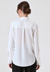 Blusa bianca a maniche lunghe con una texture liscia simile alla seta, dotata di un colletto classico e dettagli con bottoni sui polsini, con un orlo curvo.