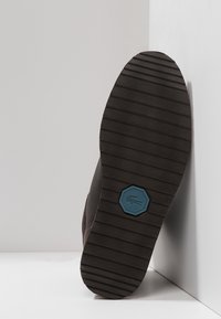 Suela de un zapato marrón con ranuras horizontales y un logo hexagonal azul con la figura de un cocodrilo, colocado sobre una esquina blanca.