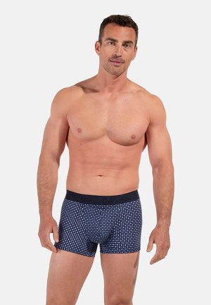 Oben ohne stehender Mann vor weißem Hintergrund, der marineblaue Boxershorts mit kleinem weiß kariertem Muster und schwarzem Bund trägt.