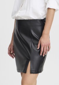 Schwarzer Leder-Minirock mit tailliertem Design, ausgestattet mit einem seitlichen Schlitz und Nähten an der Vorderseite, präsentiert an der Hüfte eines Models.