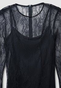 Robe en dentelle noire avec un corsage ajusté, des manches longues transparentes et un col rond. Motif floral texturé avec une fermeture éclair dissimulée dans le dos.