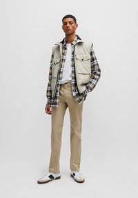 Pantalons cargo beige, t-shirt blanc, chemise à carreaux superposée sous un gilet sans manches gris clair avec poches, et baskets blanches avec accents noirs.