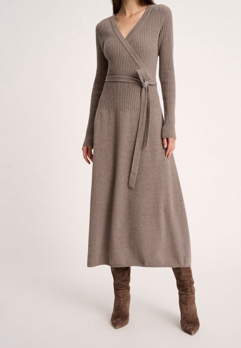 Femme porte une longue robe portefeuille en maille côtelée taupe avec une ceinture à la taille, associée à des bottes hautes en daim marron, debout devant un fond uni.