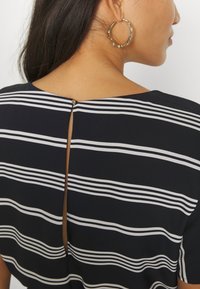 Mujer que lleva una blusa de rayas horizontales en blanco y negro con una pequeña abertura abotonada en la parte posterior del escote, y un pendiente de aro dorado texturizado.