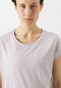 Lys lavendel kortermet topp med en glatt tekstur, rund halslinje og subtilt merke på brystet. Enkel og minimalistisk design.