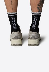 Zapatillas deportivas beige con malla texturada y acentos grises, combinadas con calcetines negros que presentan un texto blanco "NO DEJES DE EXPLORAR" y rayas.