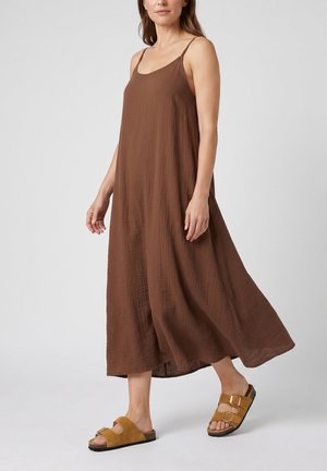 Femme marchant portant une robe midi marron ample sans manches avec de fines bretelles et des sandales marron clair à boucle, sur un fond clair uni.