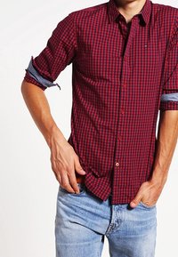 Man in een rood-zwart geblokt shirt met opgerolde mouwen, die zijn shirt in lichtblauwe jeans steekt.